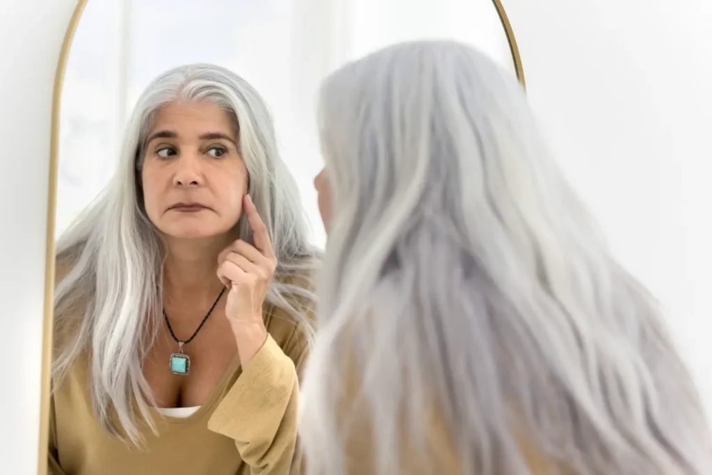 Gray-haired woman looks in a mirror while touching the skin on her cheek with one finger.